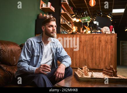 Handsome man resting after appointment in modern barbershop. Male client drinking whiskey. Bearded man sitting on leather couch in front of chessboard. Stock Photo