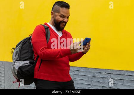 Adult Latino male (39) makes a happy gesture during a video call. He is wearing red sweater and white flannel and a backpack, yellow and gray wall bac Stock Photo