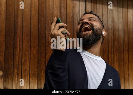 Latino adult male (39) smiles happily, holding a cell phone in his right hand. He is wearing blue sweater and white flannel, wooden slats background. Stock Photo