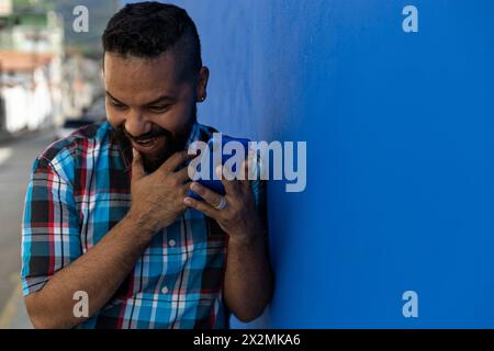 Adult male (39) with beard and plaid shirt, smiles surprised while listening to a voice message on his cell phone blue color, city background and blue Stock Photo