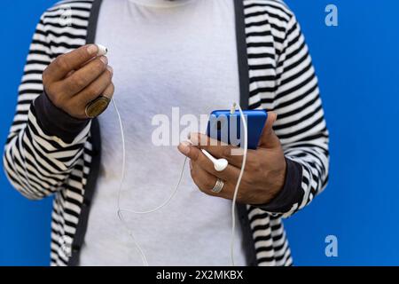 Adult male (39) with beard, short medium shot, focus on cell phone, white hands and headphones. Dressed in striped sweater, white t-shirt, blue wall b Stock Photo