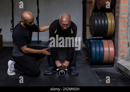 Latin American senior male with his personal trainer do rehabilitation and therapy exercises holding a kettlebell with both hands in the gym. Healthy Stock Photo