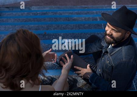 Top view of Adult man with beard and hat and young Latin American woman having relaxed conversation sitting on the stairs of a plaza. Meeting and Vaca Stock Photo