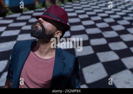 Portrait of Latin American bohemian man with beard, hat and jacket. He is wearing his hat in a funny way hiding his face. Background of shadows with r Stock Photo