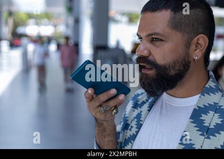 Latin American bearded man in mall spaces using his cell phone to send a voice message. Communications concept Stock Photo
