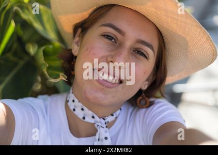 Young Latin American woman (22) with cowboy hat is sitting in the park smiling making a self-portrait with her cell phone. Concept Technology. Stock Photo