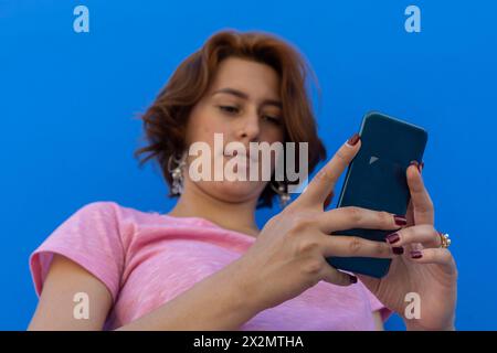 Low angle shot of Young woman (22) Latin American redhead, writing in message from her cell phone. Selective focus on cell phone. Blue wall background Stock Photo