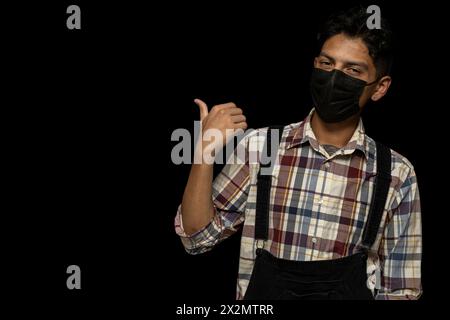 Young adult Latino male (24) with face mask points with his thumb to the right side, black background copy space. Stock Photo