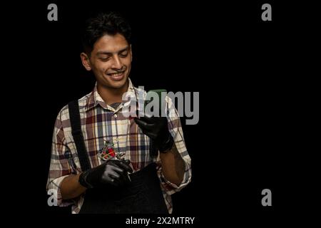 Young adult Latino male (24) holds a tattoo machine in one hand and a cell phone in the other while making a video call. Copy space with black backgro Stock Photo