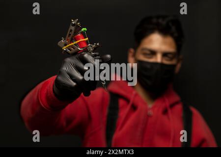 Male tattooist with mask isolated on black background showing and red sweater with selective focus on his tattoo machine. Stock Photo