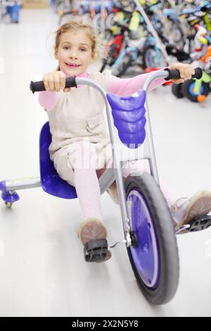 Happy girl rides blue tricycle and looks at camera in sports store. Stock Photo