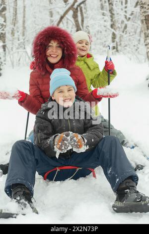 Mother, little son and daughter ride on sled in winter forest, focus on son Stock Photo