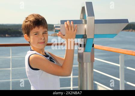 Boy stands on board of ship near big binoculars and looks at camera Stock Photo