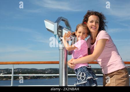 Mother and daughter stand on deck of ship near big binoculars and look at camera Stock Photo