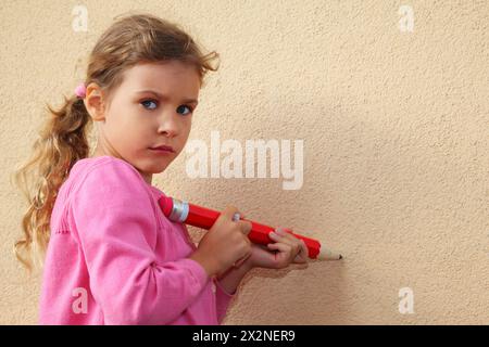 Girl paints by big red pencil on yellow wall and looks into camera. Stock Photo