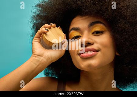 Stylish African American woman holding cream to face in trendy attire on vibrant backdrop. Stock Photo