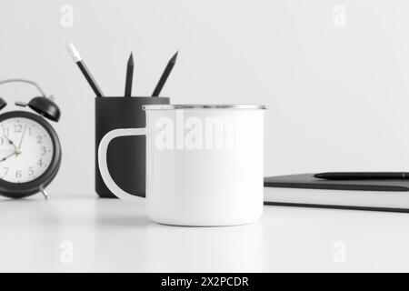 Enamel mug mockup with workspace accessories on a white table. Stock Photo