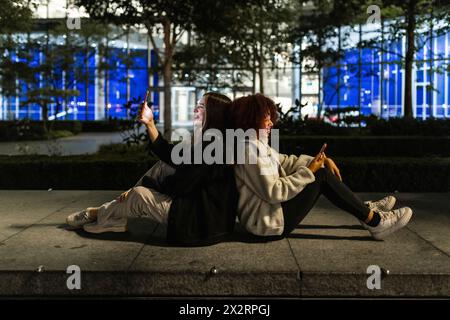 Happy friends sitting back to back and using smart phones at night Stock Photo