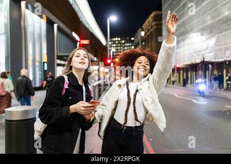 Happy woman hailing ride with friend holding smart phone on street at night Stock Photo