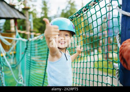 Excited smiling girl showing thumbs up gesture near net at rope park Stock Photo