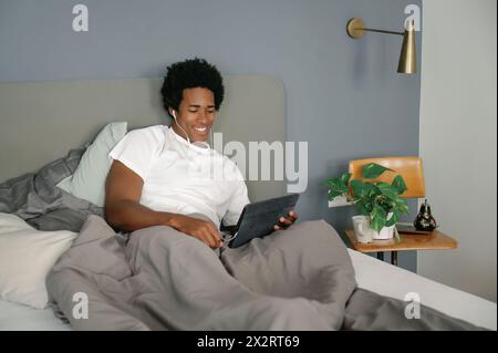 Smiling man with in-ear headphones using tablet PC on bed at home Stock Photo