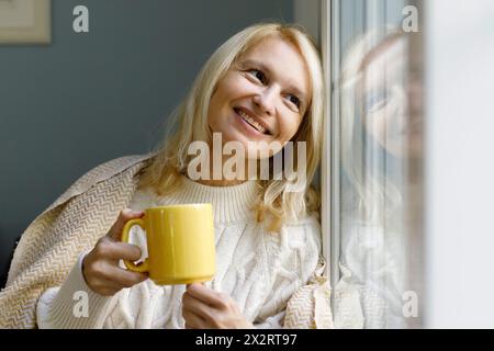 Smiling woman holding mug and looking out of window at home Stock Photo