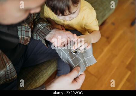 Father teaching son to sew button with needle on gray colored cloth at home Stock Photo