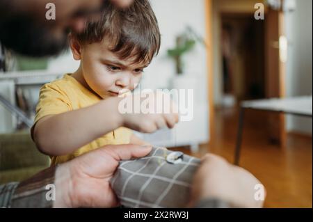 Father teaching son to sew button on gray colored cloth at home Stock Photo