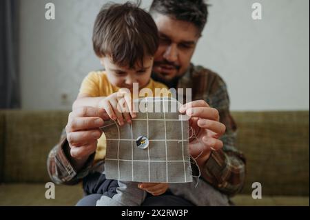 Man teaching son to sew button on gray colored cloth at home Stock Photo