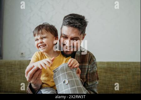 Happy father teaching son to sew button on gray colored cloth at home Stock Photo