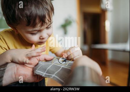 Father teaching son to sew button on gray colored cloth Stock Photo