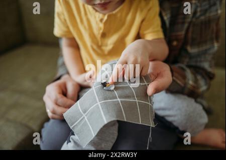Father with son learning to sew button on gray colored cloth at home Stock Photo