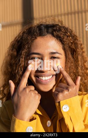 Happy beautiful young curly haired woman pointing at yellow heart shape stickers on cheeks Stock Photo