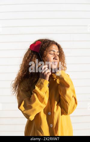 Young curly haired woman with eyes closed holding red heart shape prop in front of white shutter Stock Photo