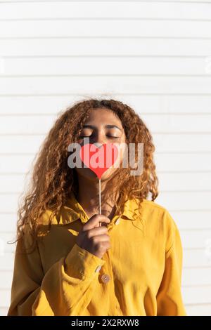 Curly haired woman with eyes closed holding red heart shape prop in front of white shutter Stock Photo