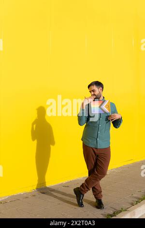 Man covering mouth and holding rainbow flag in front of yellow wall on sunny day Stock Photo