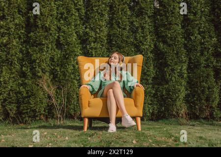 Smiling woman wearing wireless headphones and sitting on yellow armchair near trees in back yard Stock Photo