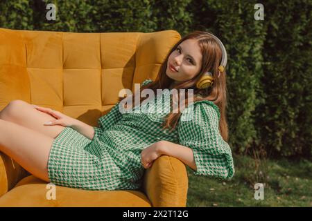 Smiling young woman wearing wireless headphones and sitting on yellow armchair Stock Photo