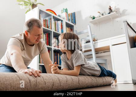 Father talking to boy lying on rolled up carpet at home Stock Photo