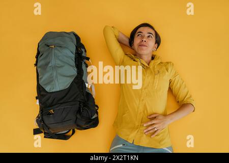 Thoughtful woman lying with backpack on yellow background Stock Photo