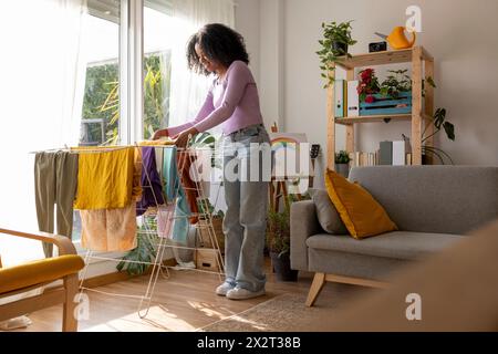 Smiling young woman drying clothes on rack by sofa in living room Stock Photo