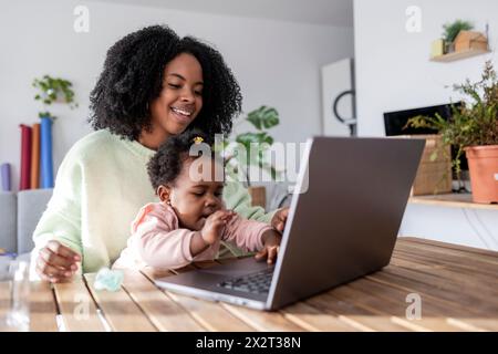 Happy working mother looking at daughter playing with laptop in home Stock Photo