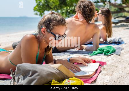 Woman reading book lying down next to friends at beach Stock Photo
