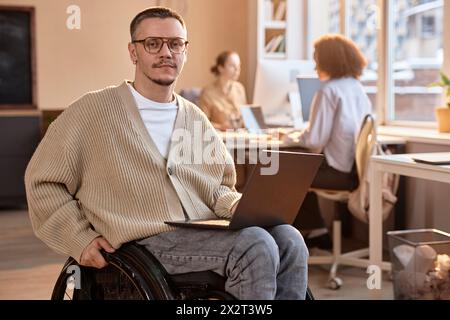 Businessman sitting with laptop in wheelchair at office Stock Photo
