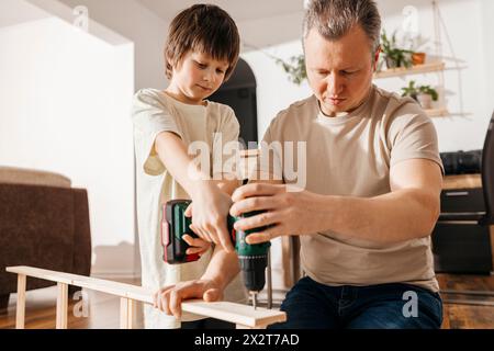 Father assisting son in using power tool for assembling wooden furniture Stock Photo