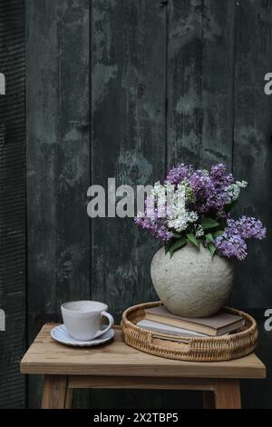 Moody farm spring breakfast still life. Purple, white lilac flowers bouquet in textured vase with cup of coffee, tea. Wicker tray, vintage books Stock Photo