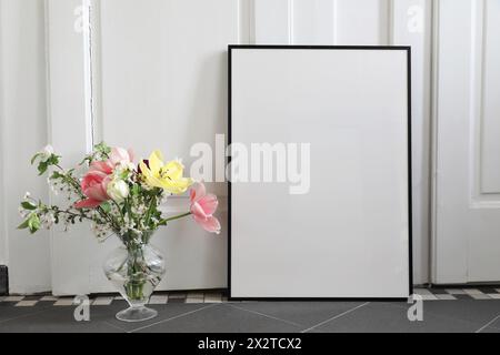 Spring still life scene. Tulips, cherry tree blossoms bouquet in glass vase. Blank black picture frame, poster mockup on grey tile floor. White wall Stock Photo