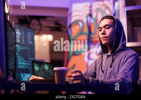 Asian hacker drinking coffee to go and hacking computer network system. Internet criminal in hood taking paper mug while coding illegal malicious software and breaking law Stock Photo