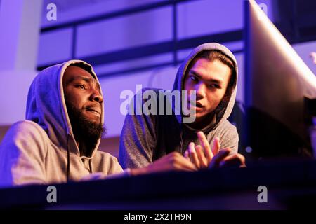 Hackers collaborating while discussing criminal activity and planning cyberattack. African american and asian criminals programming malicious software to break into database Stock Photo