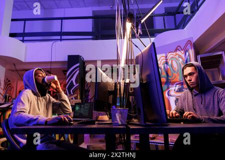Hackers breaking website and getting unauthorized access while drinking coffee. Criminals programming malicious software and planning cyberattack in abandoned warehouse at night Stock Photo